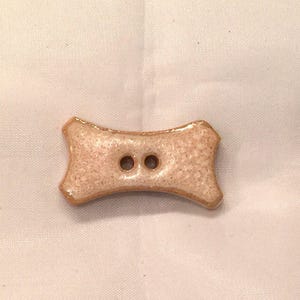 May include: A bone-shaped button with two holes. The button is a light tan color with a speckled texture and a glossy, glazed finish. The edges are a darker brown.