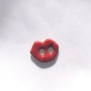 May include: A small, red ceramic button shaped like a pair of lips. The button has two holes for sewing and is set against a white background. The button is a bright, glossy red with small dark spots.