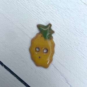 May include: A yellow and green pineapple-shaped button with two small holes. The button has a glossy finish and is set against a white wooden background. The button is approximately 1 inch in height.