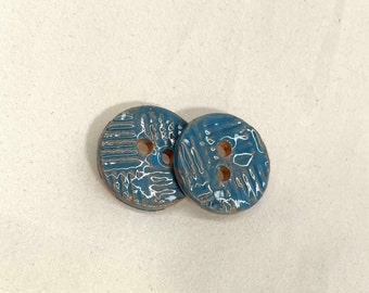 May include: Two round, ceramic buttons with a teal glaze and white accents. Each button has two holes and a textured surface with a linear pattern. The buttons are displayed on a neutral, off-white background.