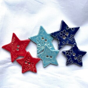 May include: A collection of star-shaped ceramic buttons in red, light blue, and dark blue. Each button features a snowflake design and two holes for sewing. The buttons are arranged on a white fabric background.