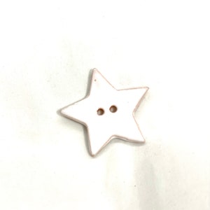 May include: A white star-shaped button with two small holes and a light brown edge. The button is centered on a white background. This decorative button is suitable for sewing and craft projects.