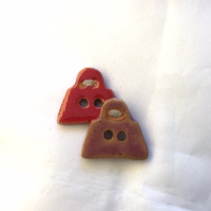 May include: Two ceramic purse-shaped buttons. One is a glossy red, the other is a muted brown. Each button has two small holes and a handle-like detail at the top. The buttons are on a white background.