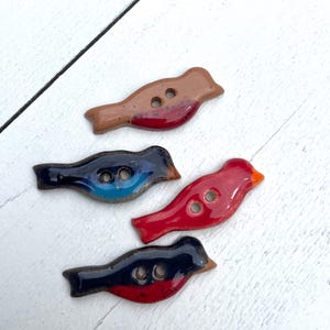 May include: Four ceramic bird-shaped buttons in various colors: brown with red, navy blue with blue, red with orange, and navy blue with red. Each button has two holes for sewing. The buttons are arranged on a white wooden surface.