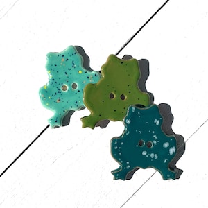May include: Three ceramic frog-shaped buttons in shades of teal, green, and dark teal. Each button has two holes for sewing and a speckled design. The buttons are arranged on a white surface.
