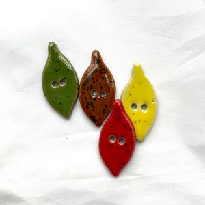May include: Four leaf-shaped ceramic buttons in shades of green, brown, red, and yellow. Each button has two holes for sewing. The buttons are arranged on a white background.