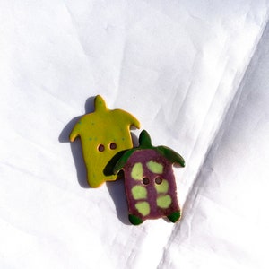 May include: Two ceramic turtle-shaped buttons. One is lime green with blue speckles, the other is purple with green accents. Both have two button holes and are set against a white background.