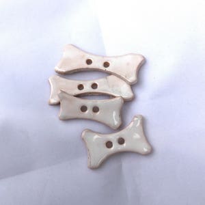 May include: Four off-white ceramic bow-tie shaped buttons with two holes each. The buttons have a glossy finish and are arranged on a white background. These decorative buttons are ideal for sewing and craft projects.