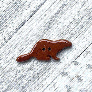 May include: A brown ceramic button in the shape of a beaver. The button has two holes and a glossy finish. The beaver button is a unique craft supply.
