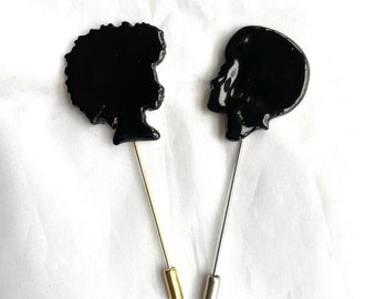 May include: Two black silhouette lapel pins. One pin features a profile of a person with an afro, the other a profile of a person with a shaved head. The afro pin has a gold-colored pin, and the shaved head pin has a silver-colored pin.
