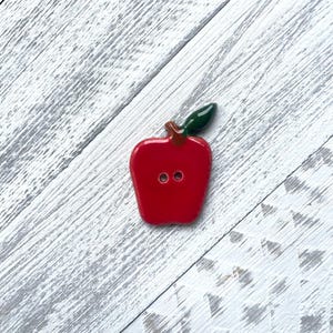 May include: A glossy red apple-shaped button with a small brown stem and a single dark green leaf attached at the top. The button features two centered holes for sewing and has a smooth, reflective surface finish throughout the entire piece.