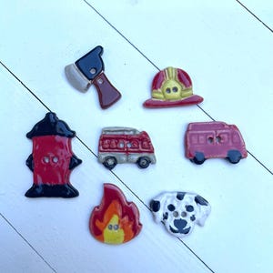 May include: A collection of decorative buttons with a firefighter theme. The buttons include a red fire hydrant, a fire axe, a firefighter's helmet, a fire truck, a Dalmatian dog, a flame, and a pink fire truck. The buttons are painted with bright colors.