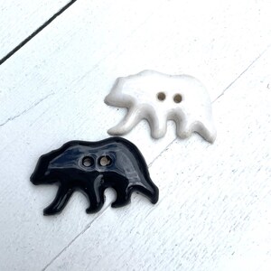 May include: Two bear-shaped buttons. One button is glossy black, and the other is white. Each button has two holes for sewing. The buttons are on a white, textured surface.