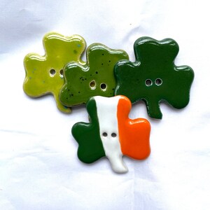 May include: Four ceramic shamrock buttons in shades of green, with a button featuring the Irish flag. The buttons have two holes for sewing. The shamrocks are glossy and have a smooth finish.