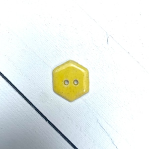 May include: A yellow, hexagonal button with two holes. The button has a slightly textured surface and is set against a white wooden background.