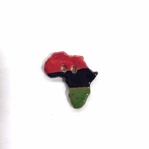 May include: A button in the shape of the African continent, painted in red, black, and green. The button has two holes for sewing. The button is on a white background.