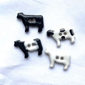 May include: Four ceramic cow-shaped buttons in black and white. Two buttons are black with white accents, and two are white with black spots. Each button has two holes for sewing. The buttons are on a white background.