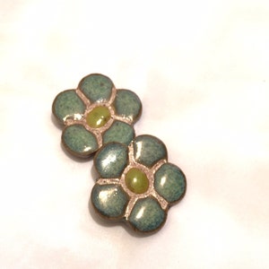 May include: Two flower-shaped earrings. Each earring has five petals in a teal-green color with a light green center. The petals are outlined in a bronze color. The earrings are on a white background.