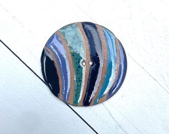 May include: A large, round button with a marbled design. The button features a mix of colors, including blue, teal, brown, and purple, arranged in vertical stripes. It has two small holes for sewing and is set against a white wooden background.