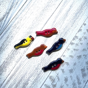 May include: Five bird-shaped buttons with two holes each, featuring glossy finishes in distinct colors including red, yellow with black accents, brown with a red breast, dark blue with light blue highlights, and black with a red breast.