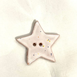 May include: A ceramic star-shaped button with two holes. The button is light pink with a white outline and small flecks of blue, yellow, and orange. The button is likely for sewing or crafting.