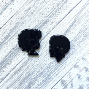 May include: Two black buttons with silhouette designs. One button features a profile of a person with an afro, and the other depicts a profile of a person with short hair. Both buttons have two holes for sewing.
