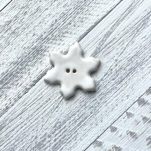 May include: A white, snowflake-shaped button with two holes for sewing. The button has a glossy finish and is set against a light gray and white wooden background. This decorative button is ideal for sewing and craft projects.