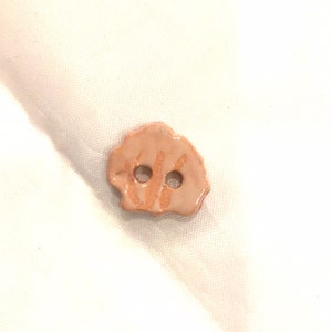 May include: A ceramic button with a light peach color and two holes. The button has a textured, irregular shape with a glossy finish. The button's surface has a subtle, hand-painted design in a darker peach hue.