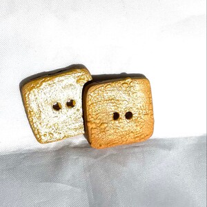 May include: Two square buttons with a textured, gold-colored surface and two holes each. The buttons have a slightly rounded shape and a dark brown edge. They are set against a white background.