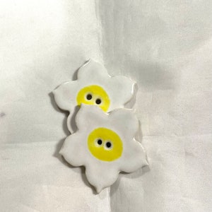 May include: Two ceramic flower-shaped buttons. Each button has a white petal design with a yellow center and two black button holes. The buttons are on a white background.