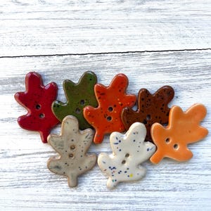May include: A collection of seven ceramic buttons shaped like oak leaves, each featuring two center holes. The buttons are finished in various glossy colors including red, green, orange, brown, beige, and white, with speckled details.