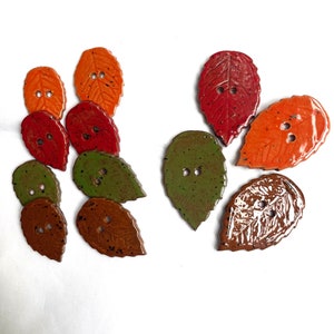 May include: A collection of leaf-shaped buttons in various colors, including orange, red, green, and brown. Each button has two holes for sewing and a glossy finish. The buttons are arranged on a white surface.