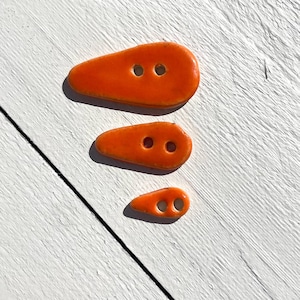 May include: Three orange, teardrop-shaped buttons with two holes each. The buttons are arranged in descending order of size, with the largest at the top and the smallest at the bottom. The buttons are on a white wooden surface.