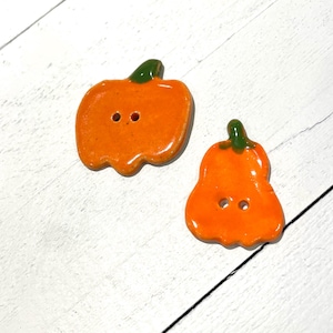 May include: Two orange pumpkin-shaped buttons with green stems. Each button has two holes for sewing. The buttons are glossy and appear to be ceramic or a similar material. The background is a white wooden surface.