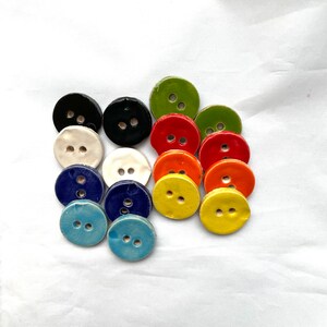 May include: A collection of colorful ceramic buttons in various shades, including black, white, green, red, orange, yellow, and blue. Each button is round with two holes for sewing. The buttons are arranged on a white surface.