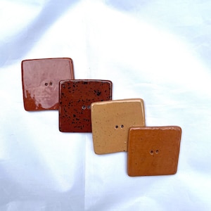 May include: Four square ceramic buttons in shades of brown, ranging from a glossy reddish-brown to a speckled dark brown. Each button has two holes for sewing. The buttons are arranged diagonally.