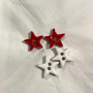 May include: Four star-shaped buttons. Two are red, and two are white. Each button has two holes for sewing. The buttons are on a white, textured surface.