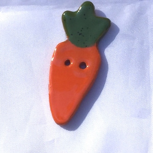 May include: A ceramic carrot-shaped button with an orange body and green top. The button has two holes for sewing. The carrot button is a whimsical and decorative element for sewing projects.