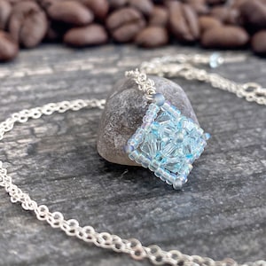 May include: A delicate silver chain necklace with a light blue crystal pendant. The pendant is a square shape and is made of small, faceted crystals.