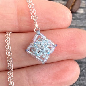 May include: A delicate silver chain necklace with a light blue and silver beaded pendant in the shape of a square.