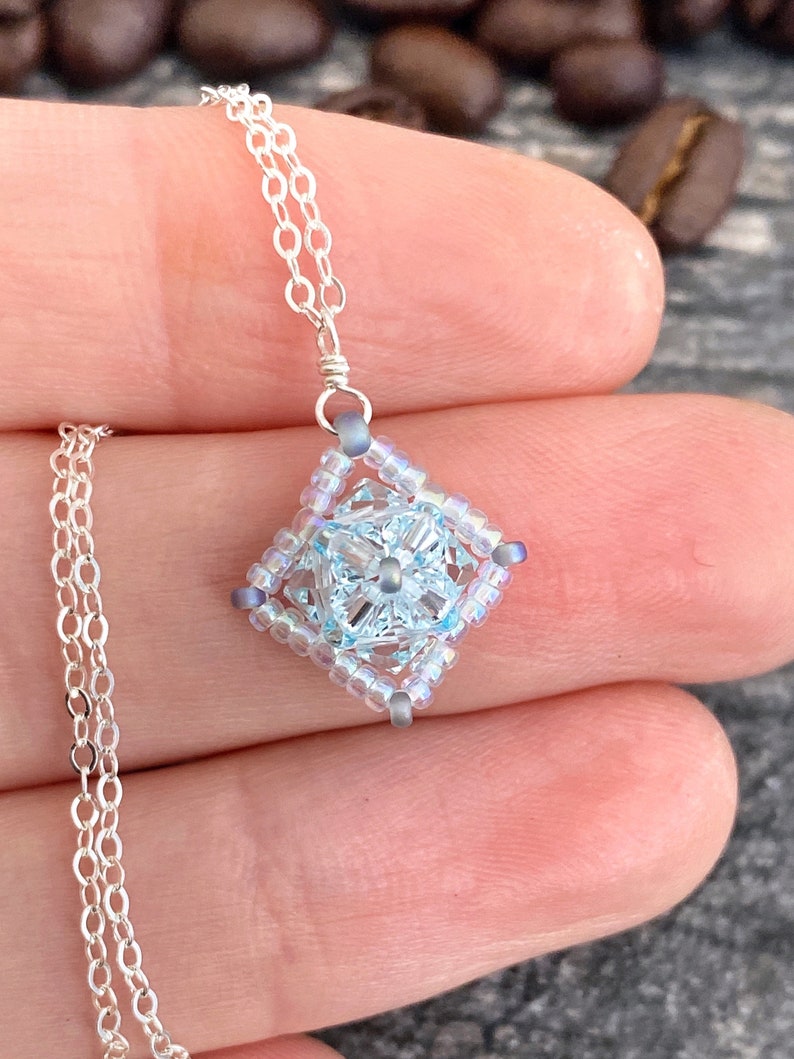 May include: A delicate silver chain necklace with a light blue and silver beaded pendant in the shape of a square.