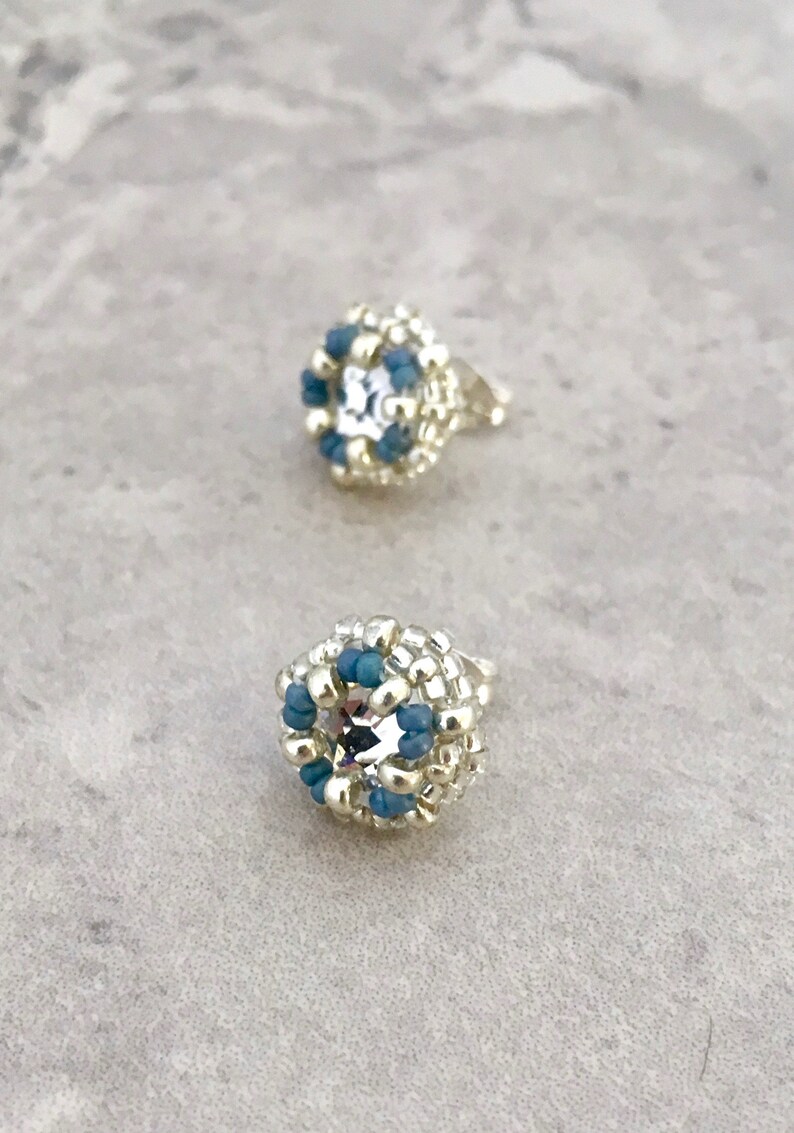 Puede incluir: Un par de pendientes de bot&oacute;n plateados con un dise&ntilde;o de cuentas azules y plateadas. Cada pendiente tiene un cristal transparente en el centro.