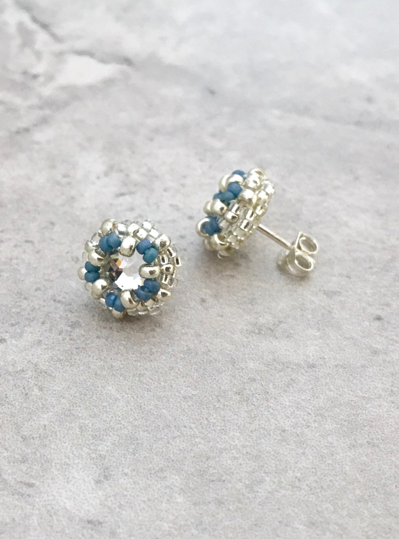 Puede incluir: Un par de pendientes de bot&oacute;n de plata con un dise&ntilde;o de cuentas azules y plateadas. Los pendientes tienen un cristal transparente en el centro del dise&ntilde;o.