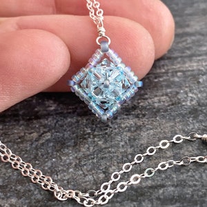 May include: A silver chain necklace with a light blue beaded pendant in the shape of a square with a flower design in the center.