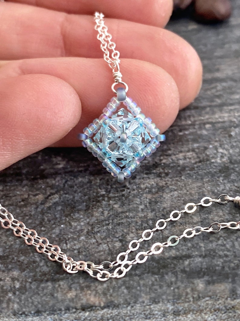 May include: A silver chain necklace with a light blue beaded pendant in the shape of a square with a flower design in the center.