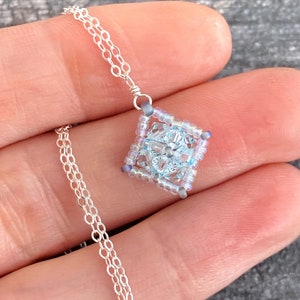 May include: A delicate silver chain necklace with a square pendant made of light blue and iridescent beads. The pendant features a clear crystal in the center.