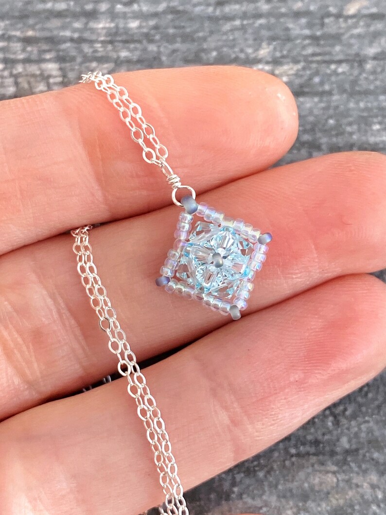 May include: A delicate silver chain necklace with a square pendant made of light blue and iridescent beads. The pendant features a clear crystal in the center.