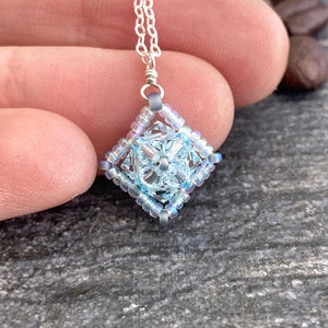 May include: A delicate silver chain necklace with a light blue and iridescent crystal pendant. The pendant is a square shape with a smaller crystal in the center.