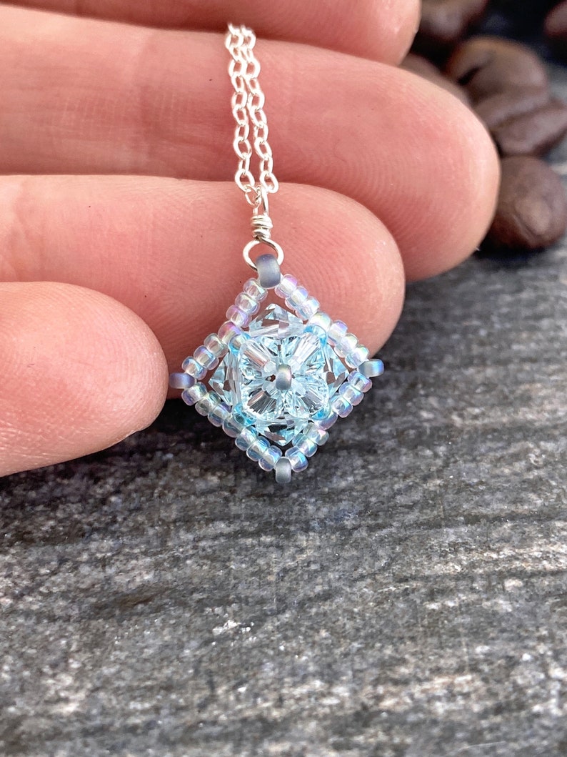 May include: A delicate silver chain necklace with a light blue and iridescent crystal pendant. The pendant is a square shape with a smaller crystal in the center.