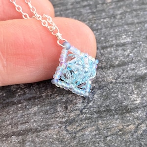 May include: A delicate silver chain necklace with a light blue and iridescent beaded pendant in the shape of a square.